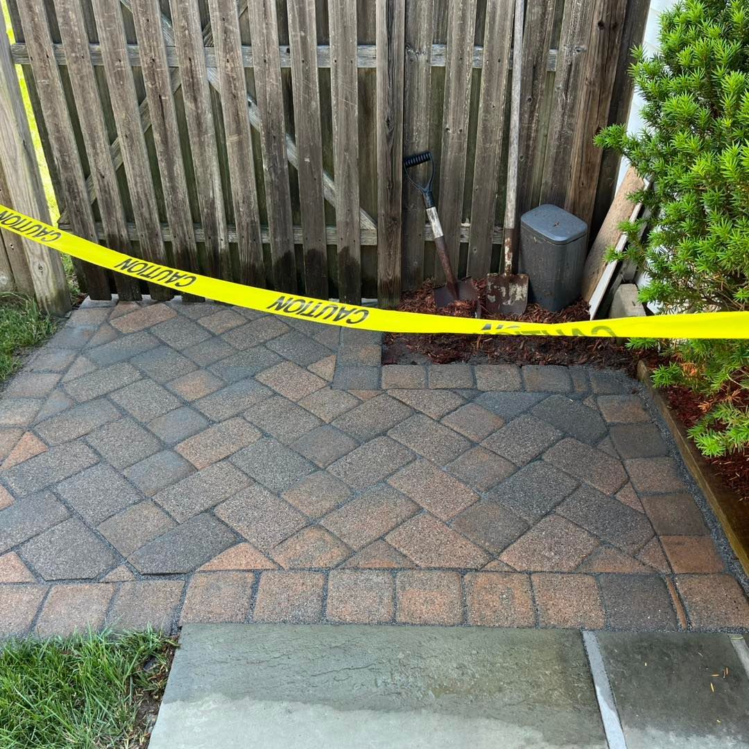 Brick patio with caution tape near a wooden fence and small evergreen bush.