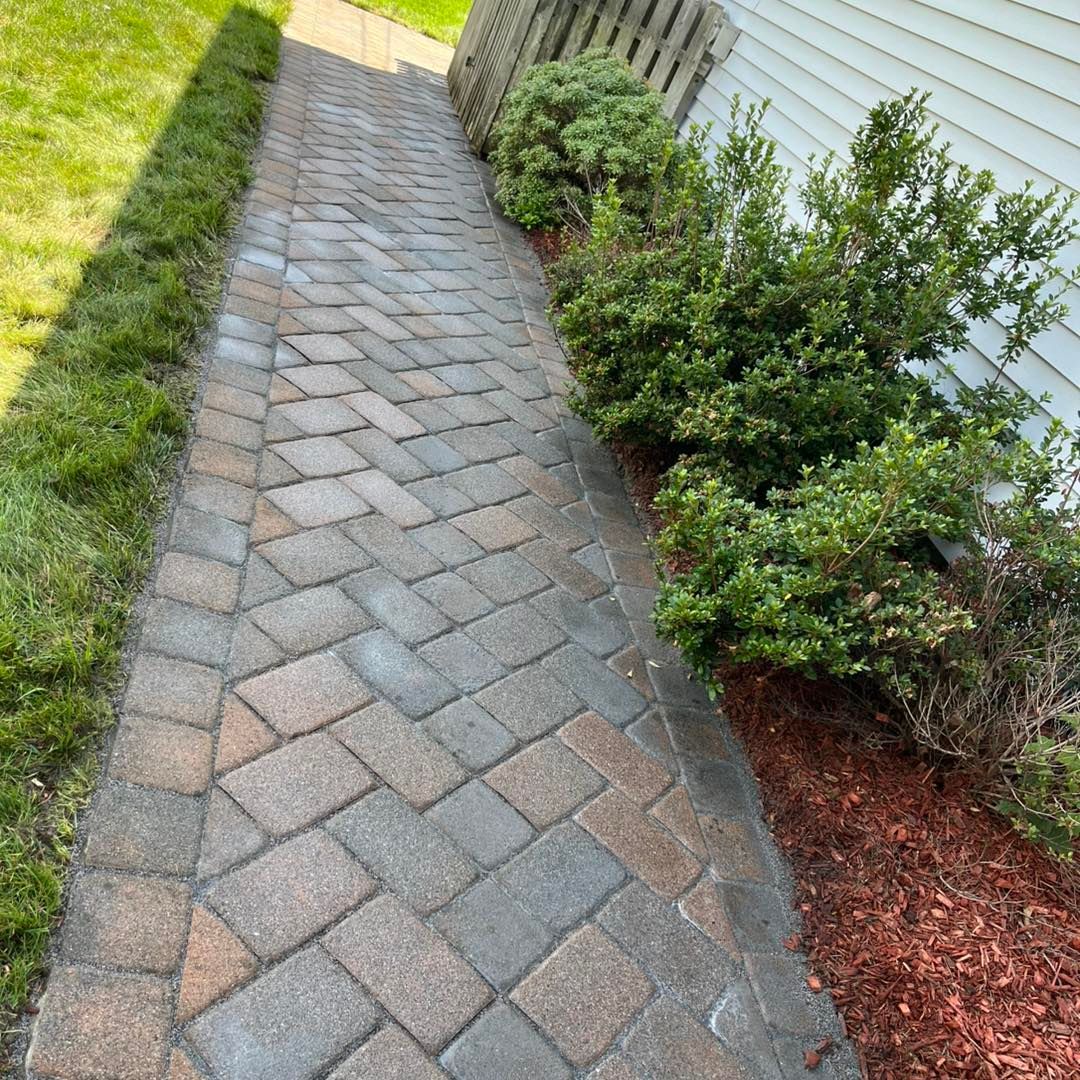 Brick walkway bordered by green grass and shrubs, leading alongside a white house.