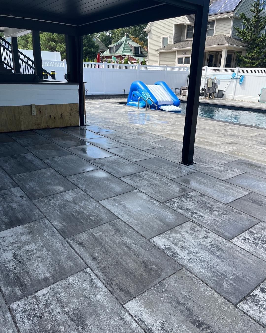 Patio with grey stone pavers, black pergola, and a pool with a blue slide.