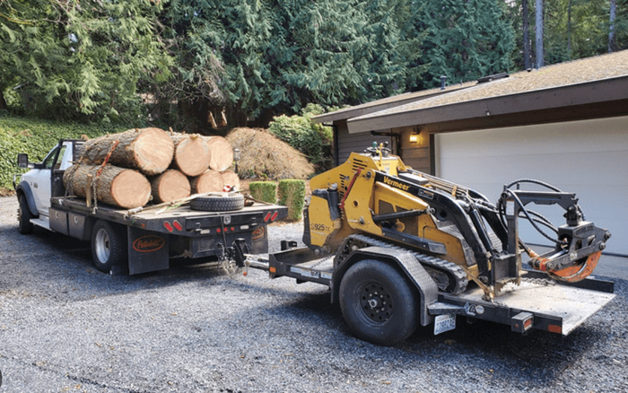 Truck hauling logs, pulling a trailer with a yellow stump grinder, parked in front of a house.