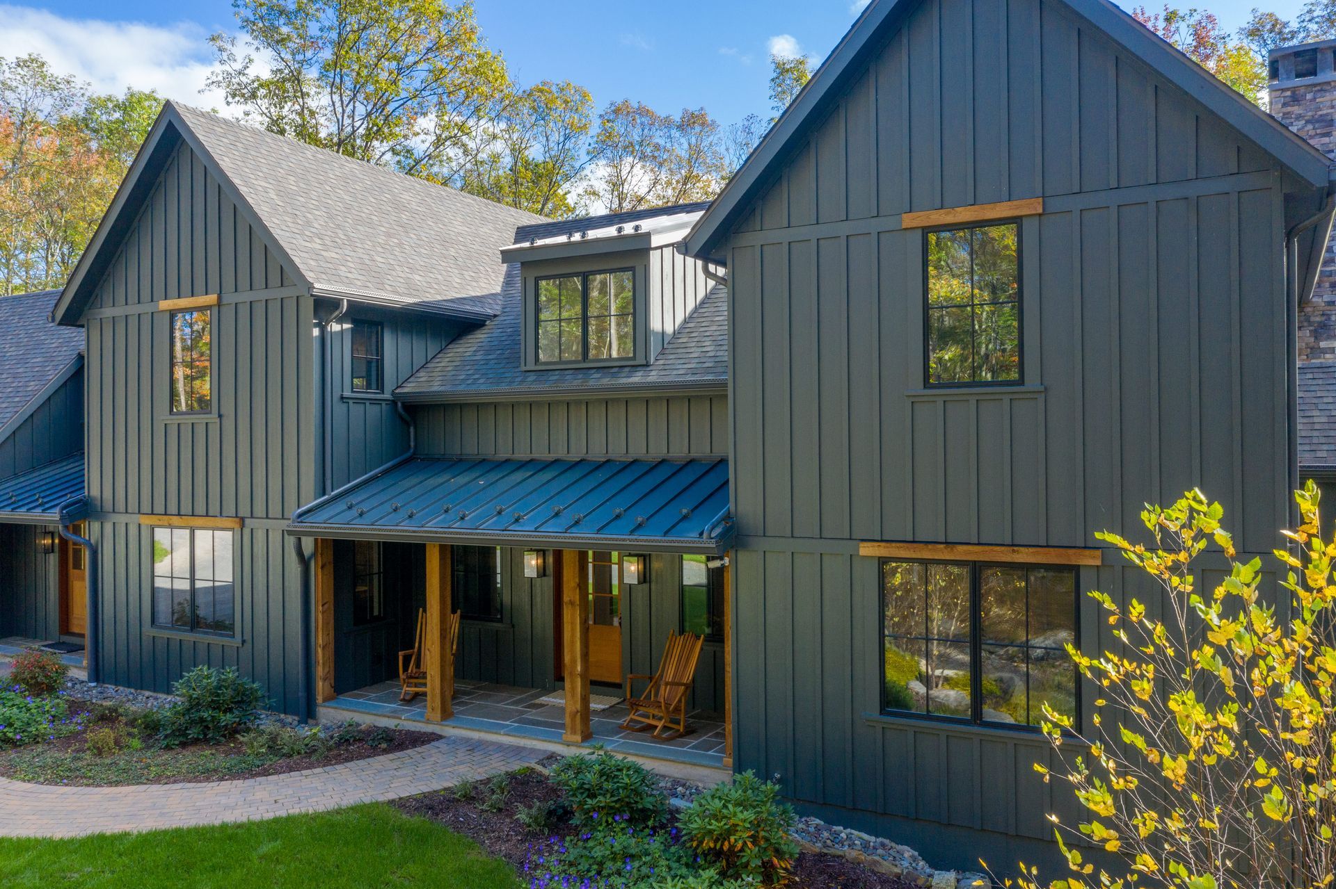 A modern two-story home with dark gray vertical siding, a metal porch roof, and wooden accents, surrounded by autumn trees.