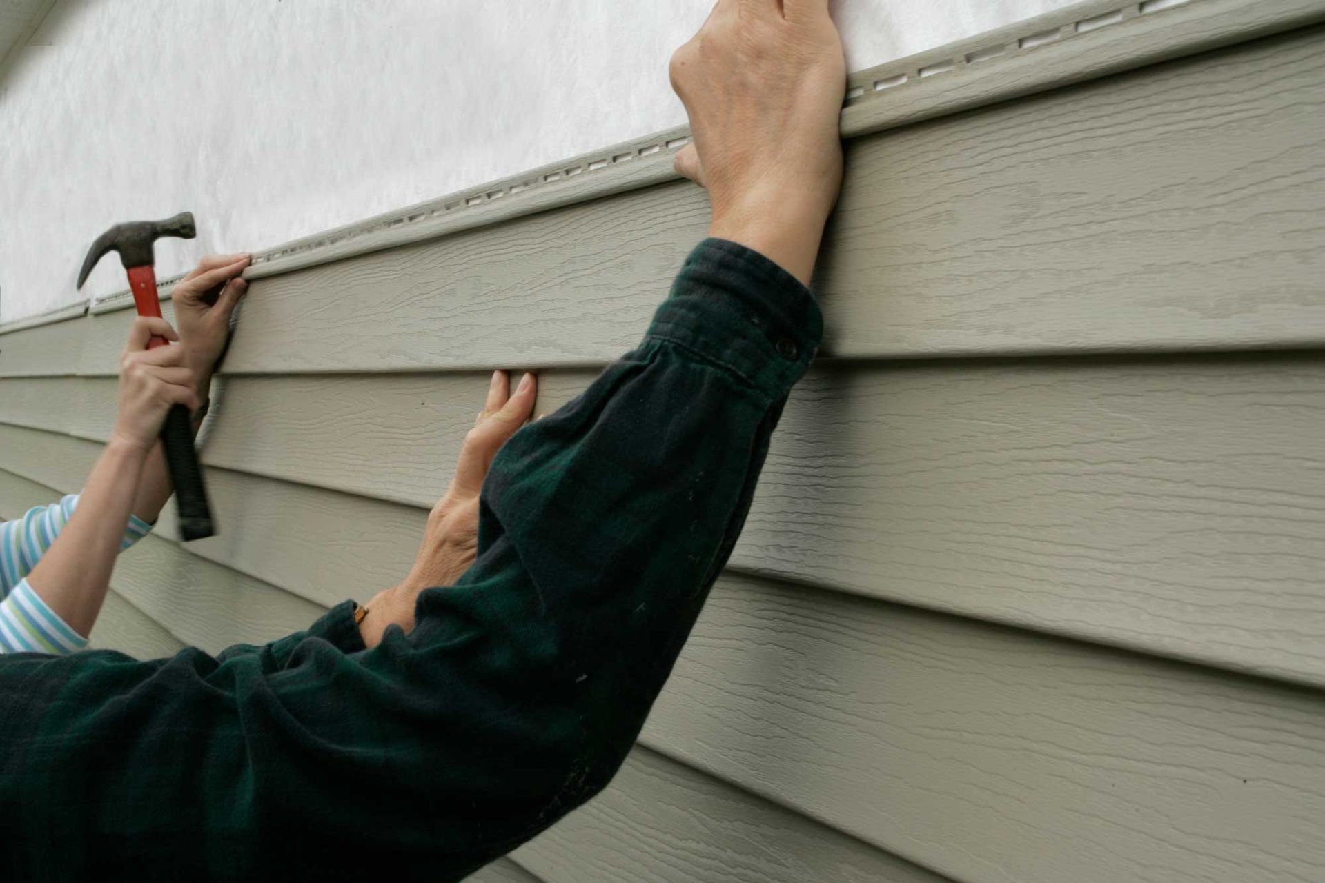 Hands installing tan siding on a wall with a hammer.