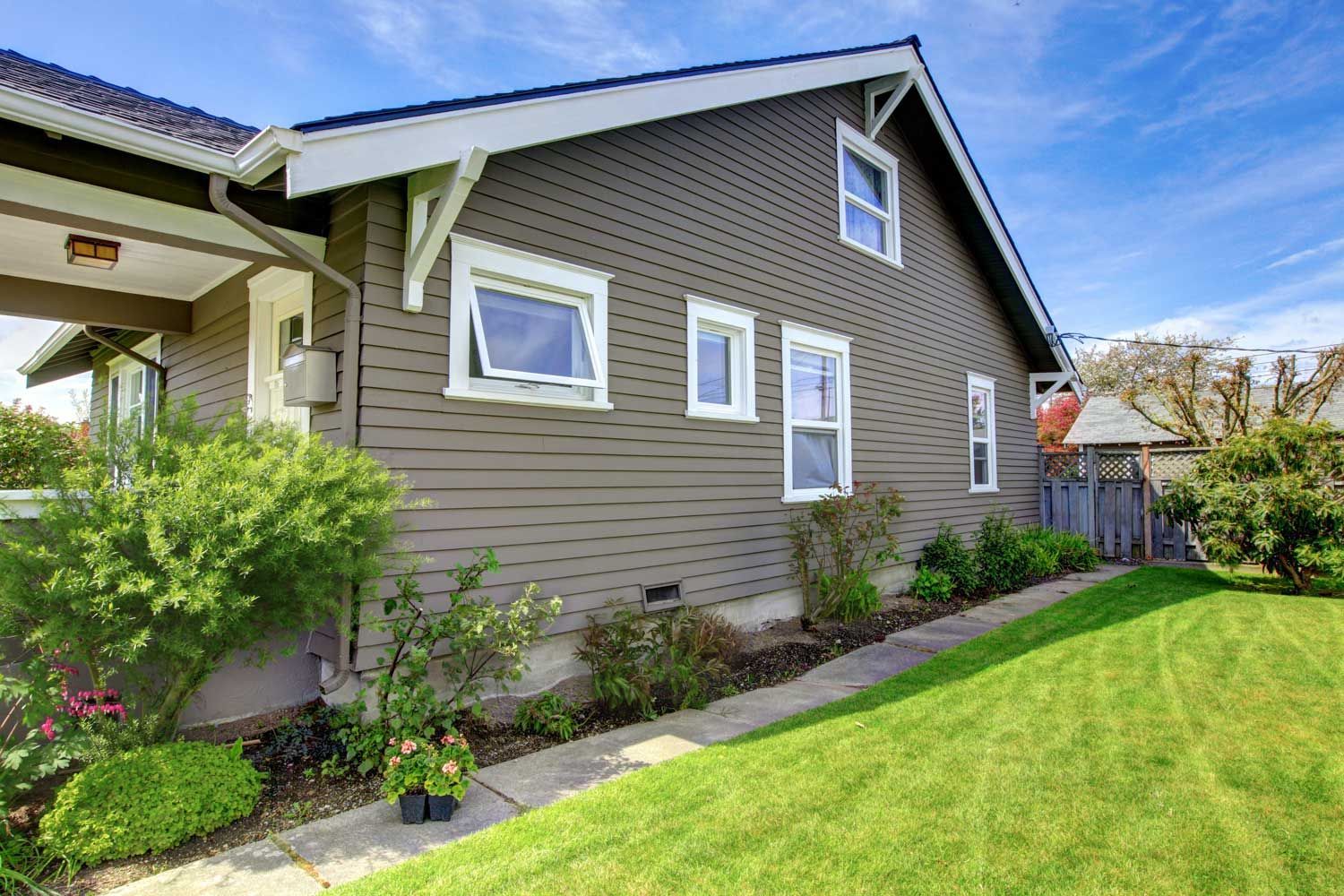 Brown house with white trim, green lawn, and shrubs under a blue sky.