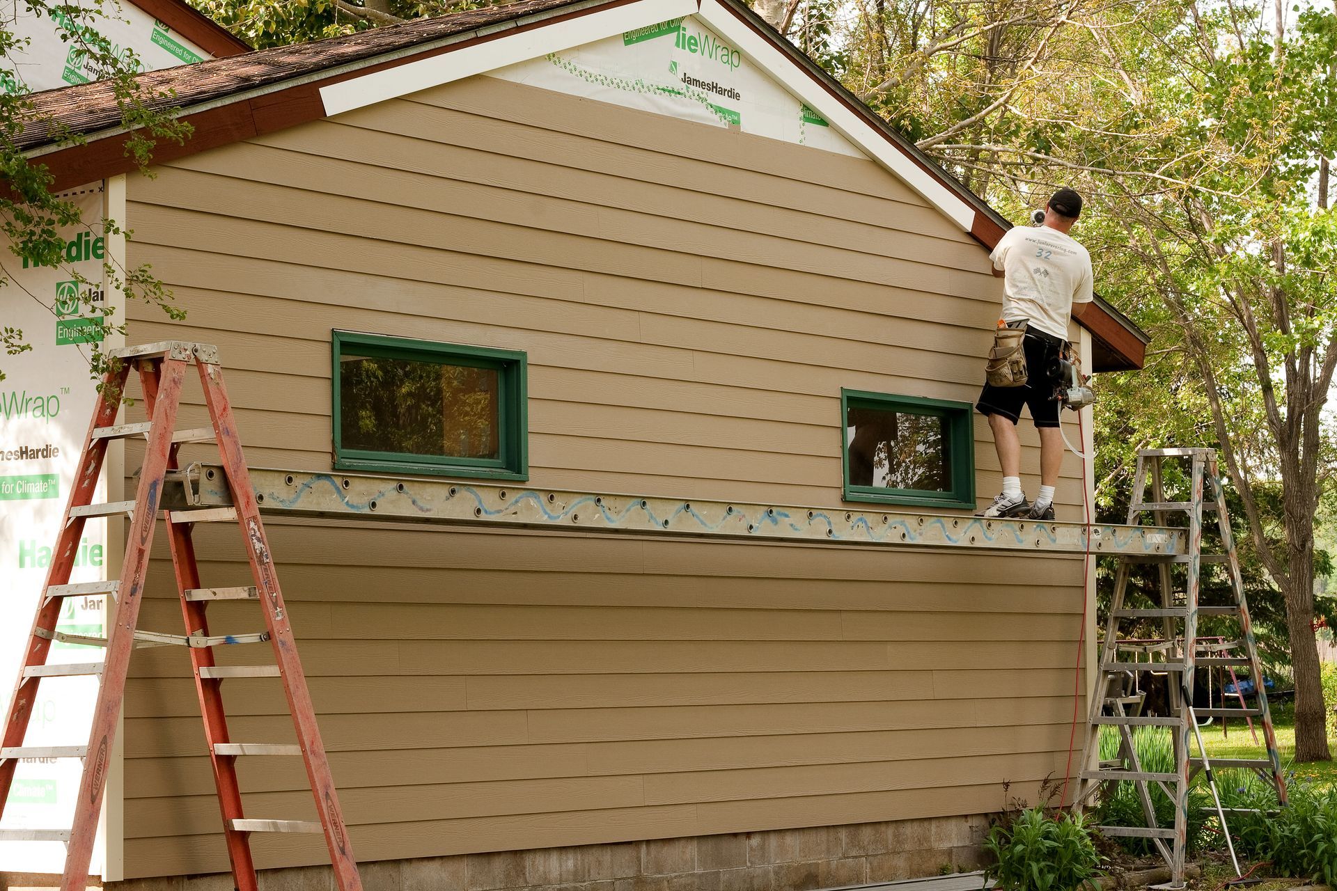 A worker installs beige lap siding on the side of a house while standing on a metal scaffold supported by two ladders.