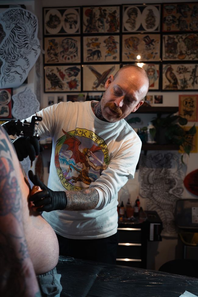 Tattoo artist working on a client’s arm in a studio, with tattoo design sheets on the wall.