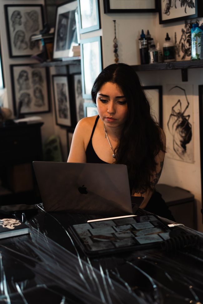 Woman working on a laptop at a desk in a softly lit room with framed artwork and shelves behind her
