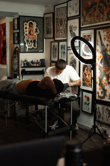 Tattoo artist working on a client in a studio with framed art and a ring light