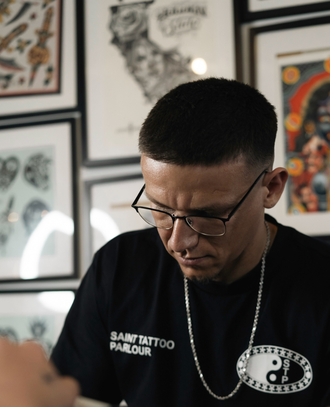 Person in glasses and black shirt looking down in a tattoo studio with framed artwork on the wall.