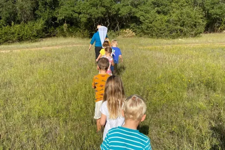 Children walking single file through a grassy field with a blue kite or banner ahead