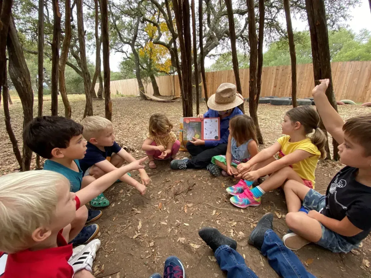 Children sitting in a circle outdoors, listening to a teacher reading from a tablet in a wooded area