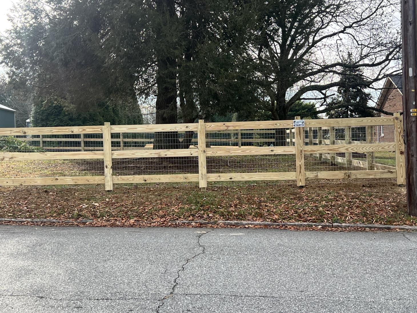 Wooden split-rail fence with wire mesh in front of trees and a house, seen from a paved road.