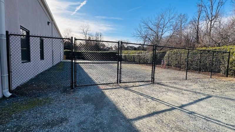 Black chain-link fence with double gates in front of a white building on a gravel lot; sunny day.