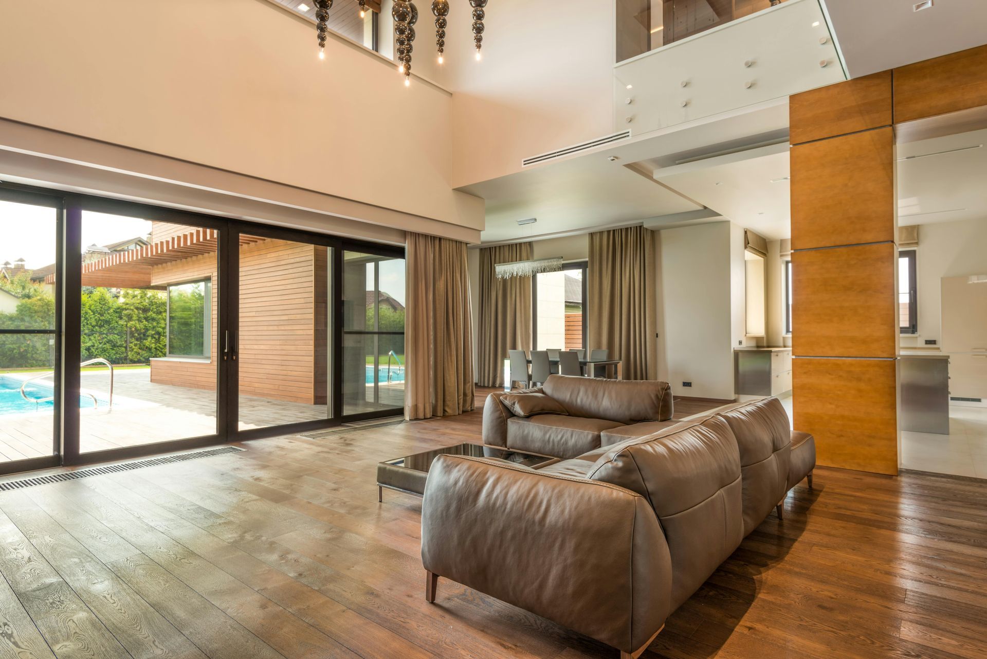 Living room with large windows overlooking a pool, brown leather couch, and wooden floors.