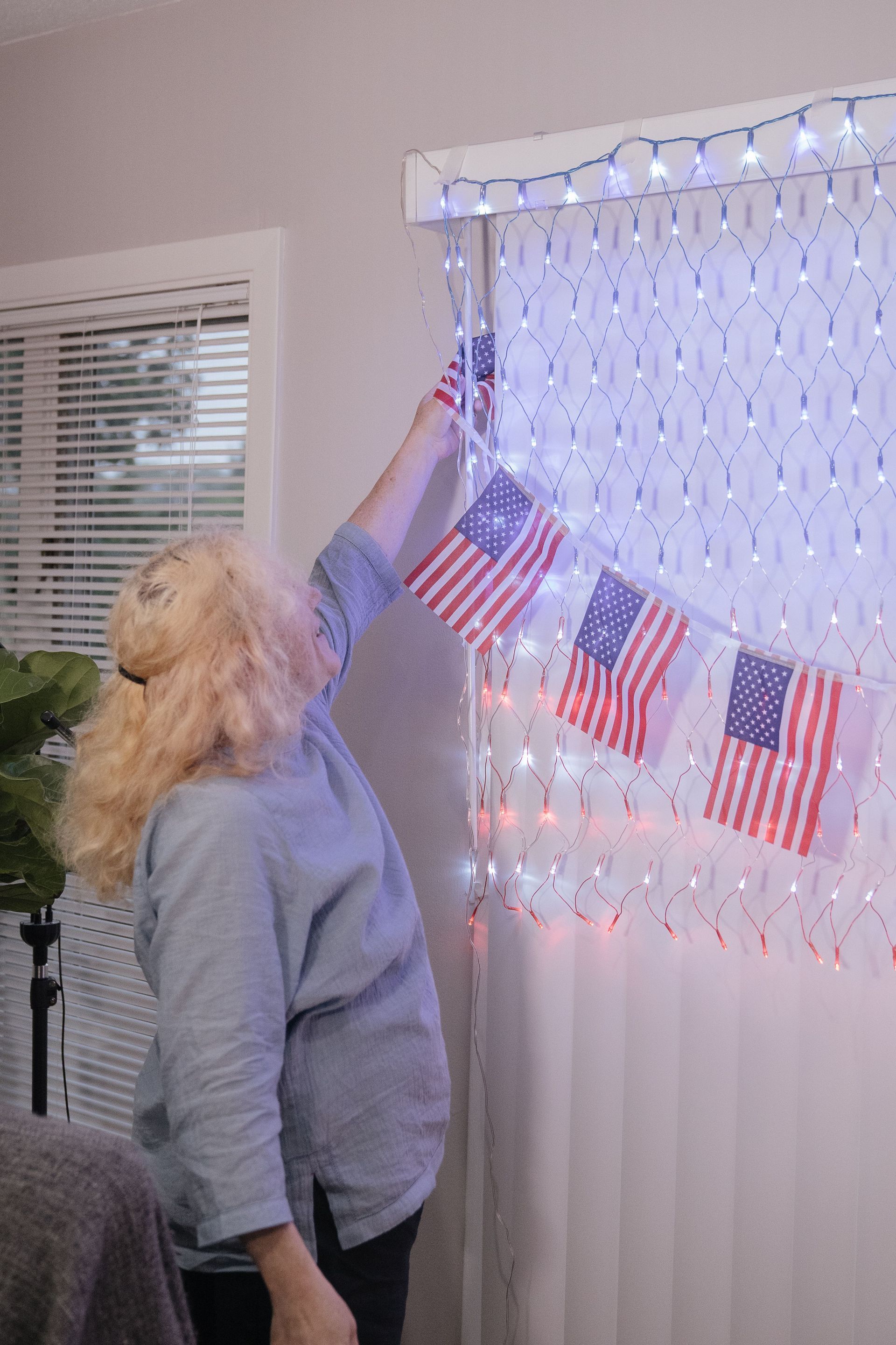 Person reaching up to hang a string of red, white, and blue flags on a lit window curtain