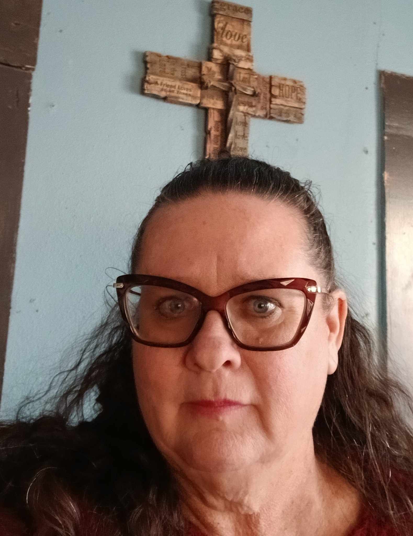 A woman wearing glasses is standing in front of a cross on a wall.