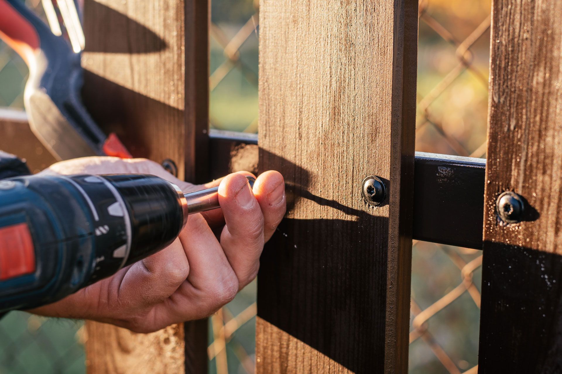 Hands using a cordless drill to screw a black metal hinge into a wooden fence post