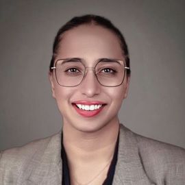 Team member Rukaiya, with glasses, smiles, wearing a gray blazer, dark top, and a gray background.