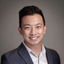 Team member Aaron, in gray suit and light shirt smiles at the camera against a gray background.