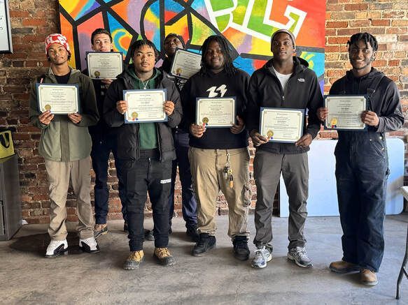 A group of men standing in front of a brick wall that's been painted. Each man is holding a certificate.
