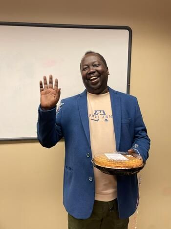 A man smiling and waving while holding a pie. He is wearing a blue professional blazer.
