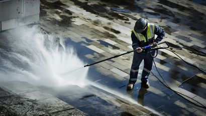 Worker pressure-washing a sloped rooftop, spraying water across dark shingles and debris