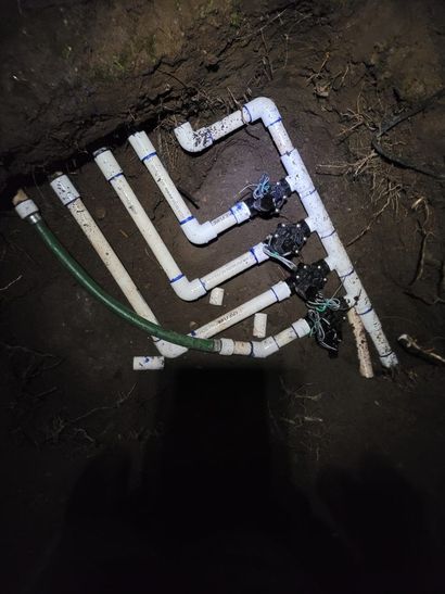 White PVC pipe network forming a shape on dirt ground, with dark fittings and a green hose nearby.
