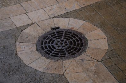 Circular drain cover set in a stone pavement with a beige tiled border.