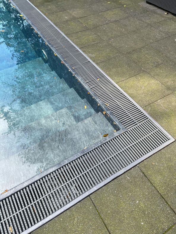 Pool corner with blue water, metal drainage grates, and tan pavement.