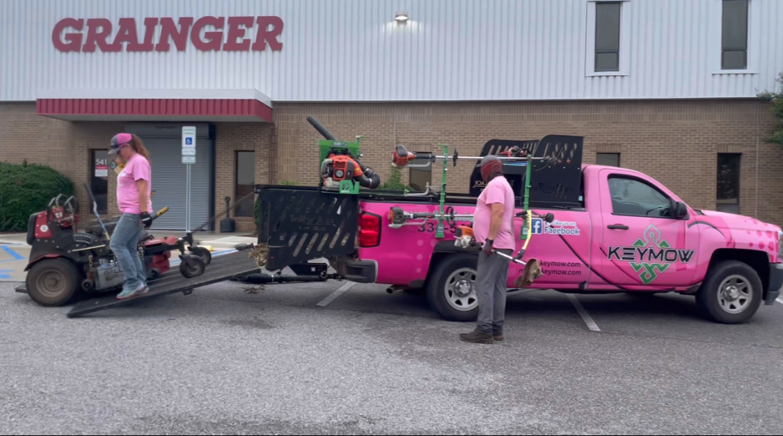 Workers grabbing lawn care equipment from a pink truck is parked in front of a building that says grainger