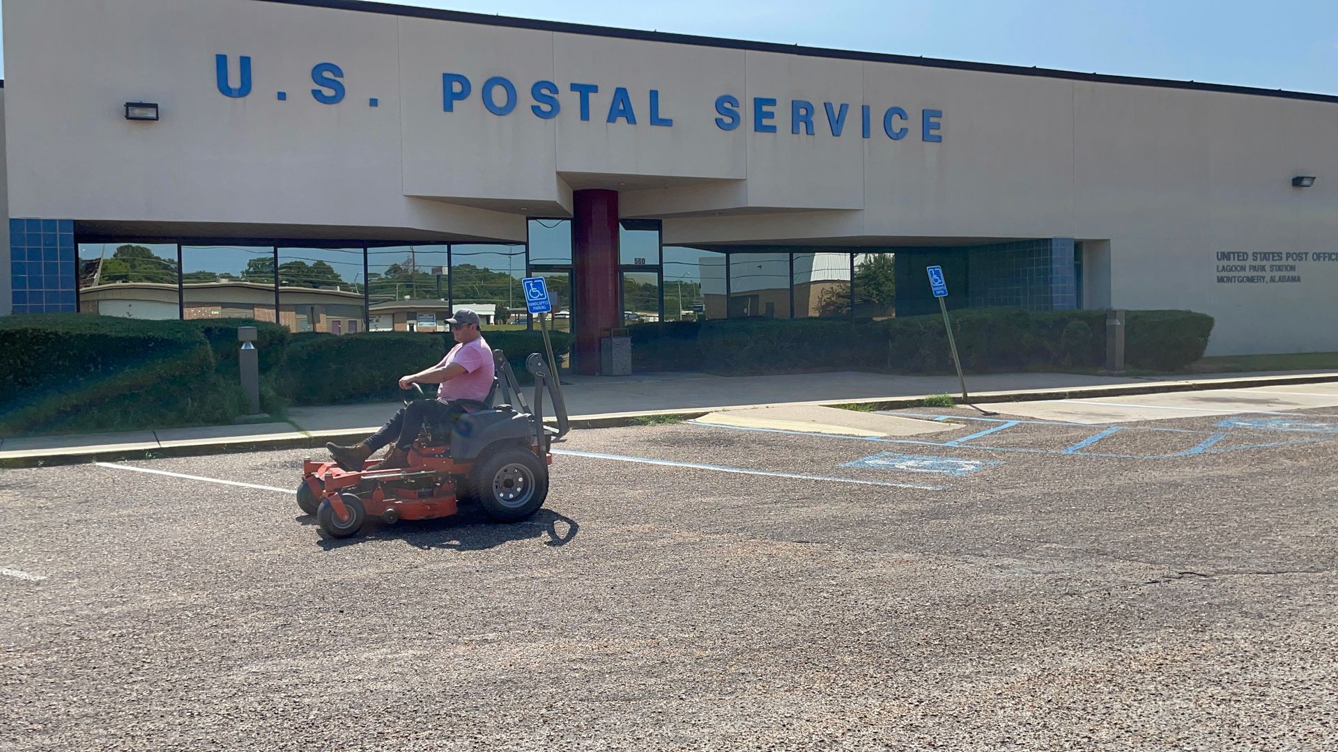 A man is riding a lawn mower in front of a u.s. postal service building.