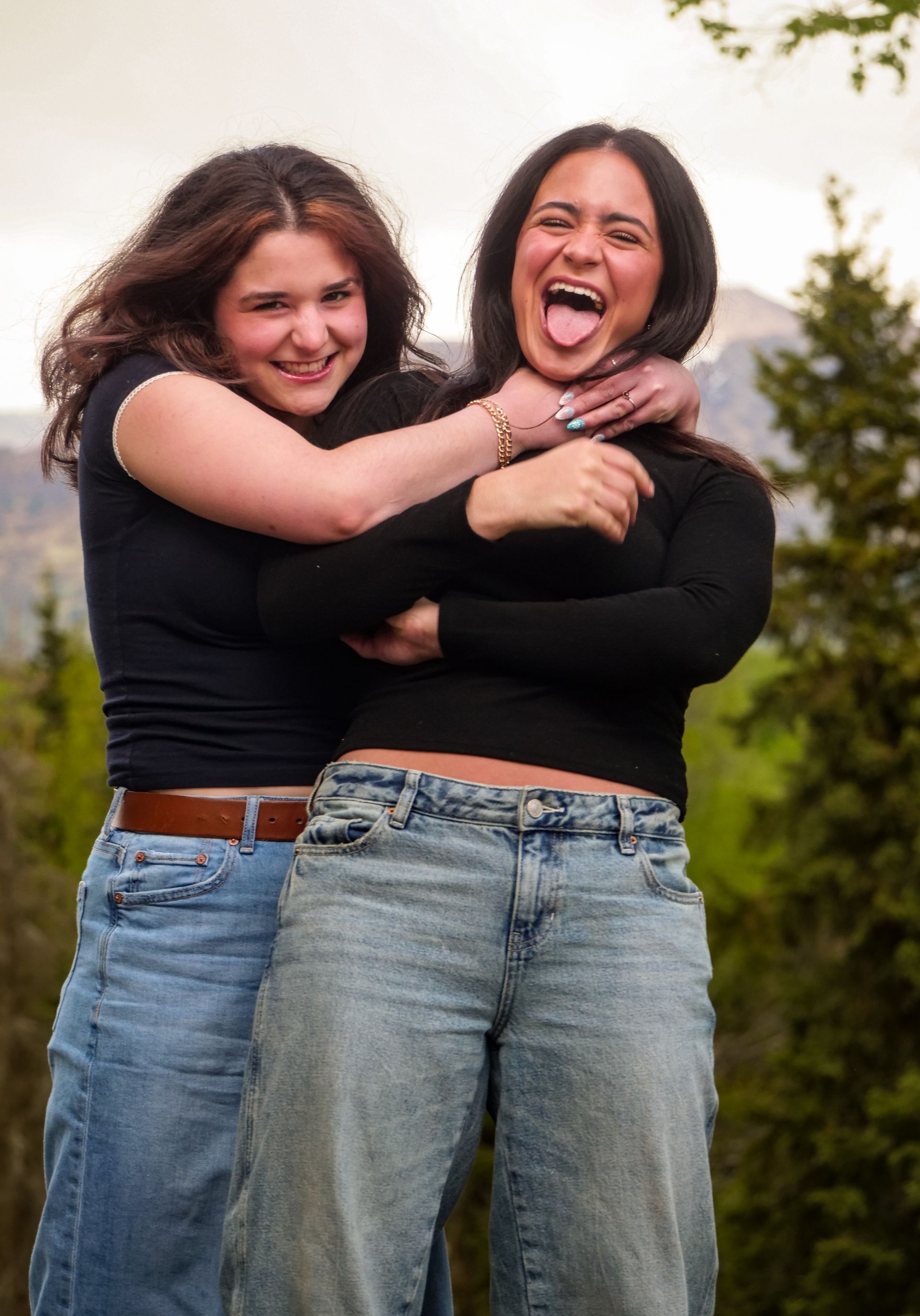 Two young women smiling and laughing; one hugs the other from behind, outdoor setting.