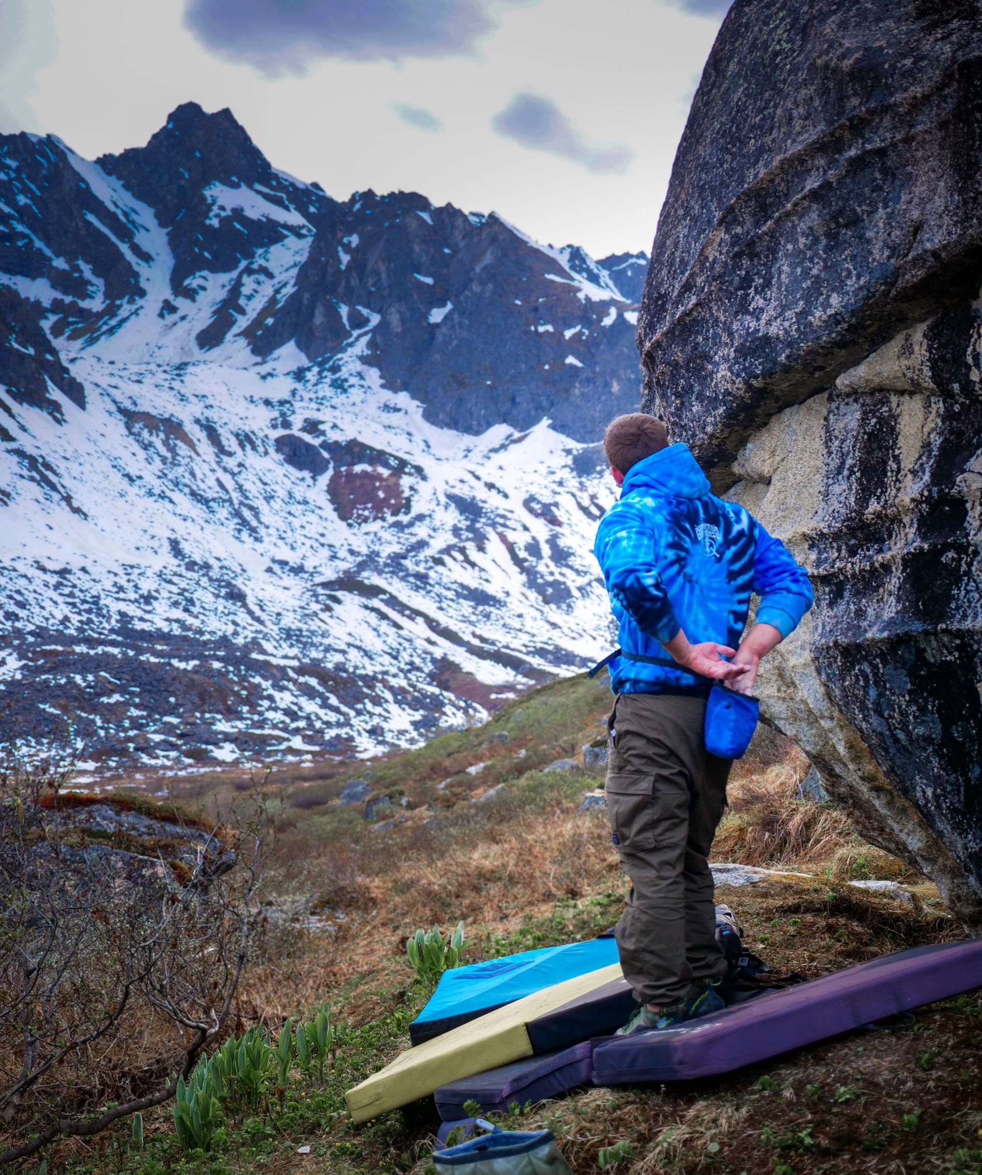 A person in blue jacket prepares to boulder outdoors, looking towards snow-covered mountains.