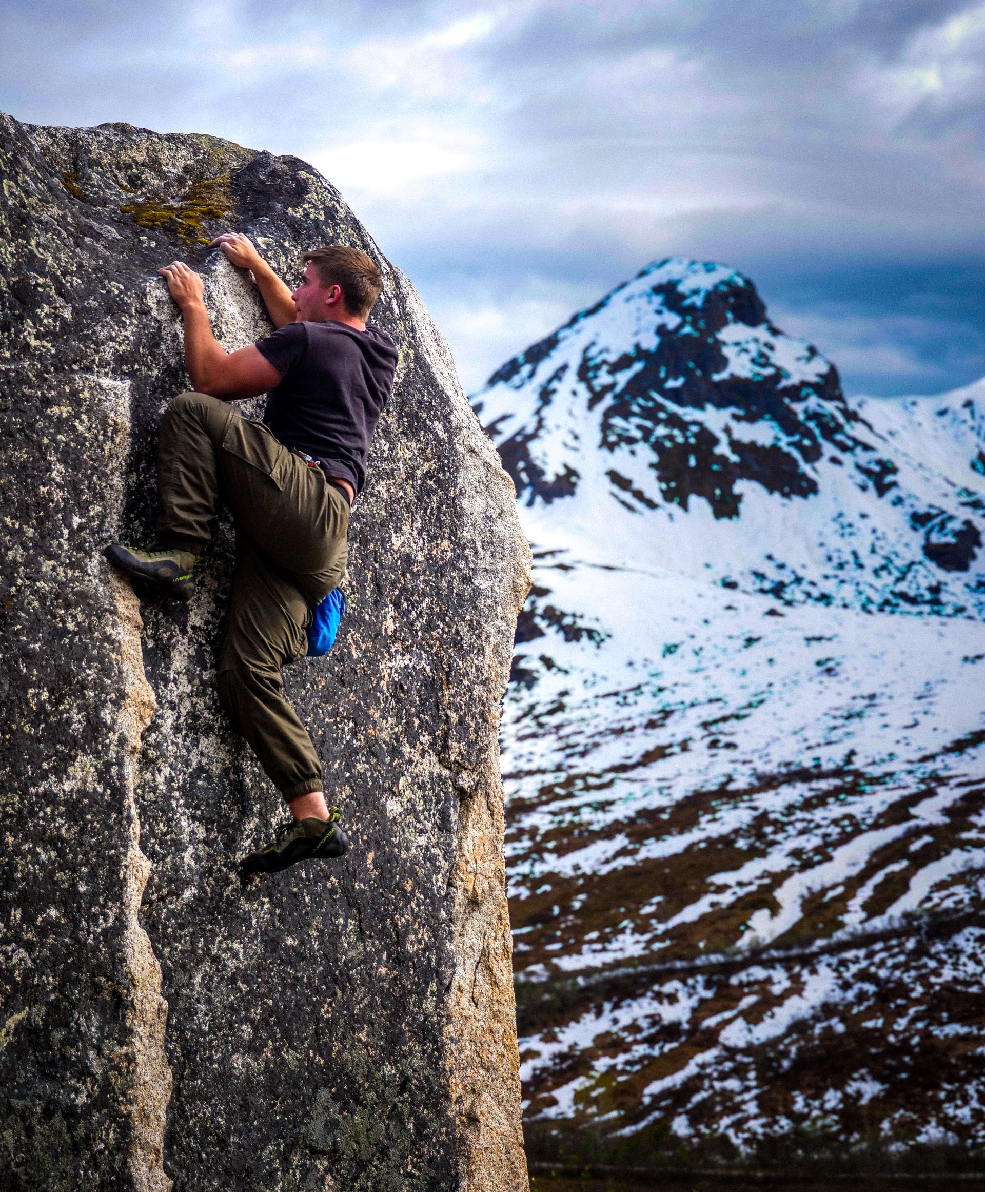 Rock climber on a boulder, snowy mountain backdrop. Dark clothes, reaching for holds.