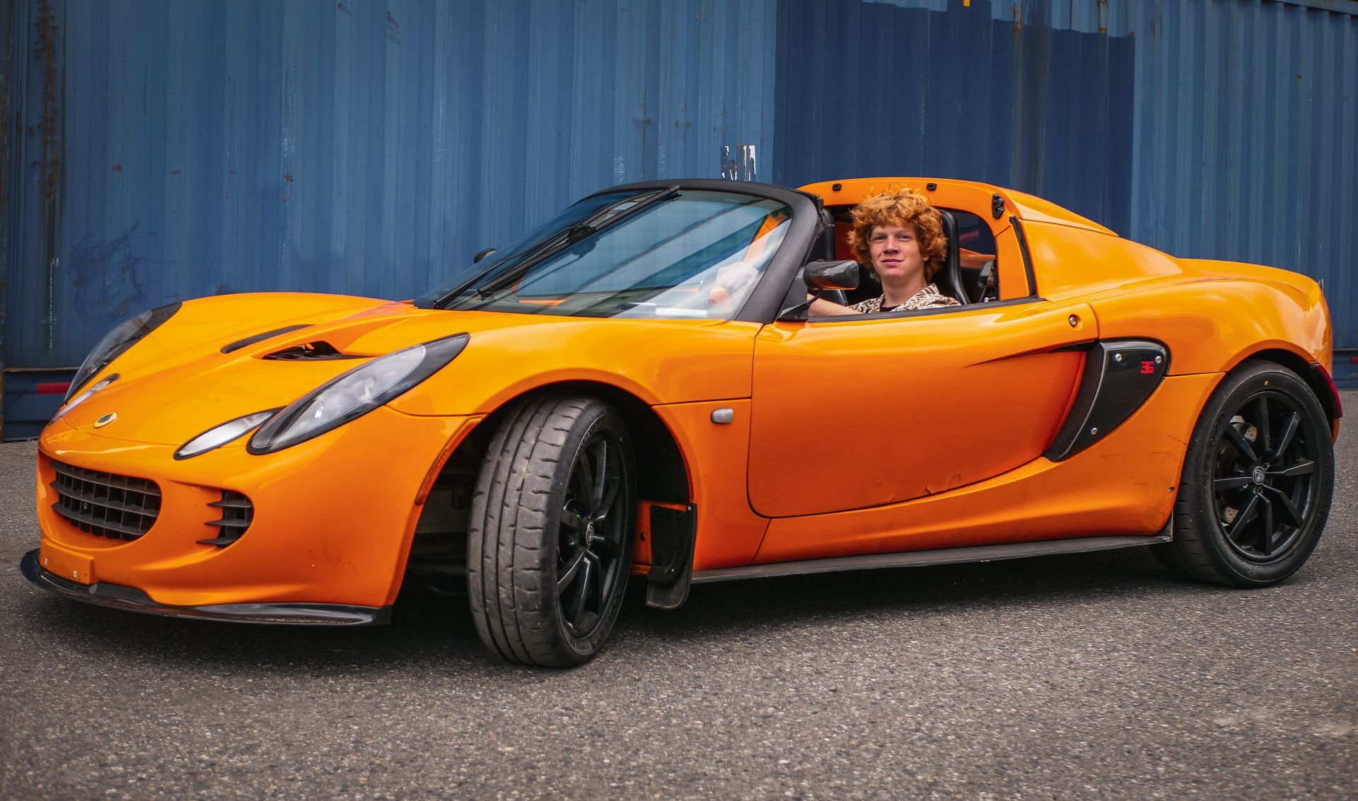 Orange Lotus sports car with a person in the driver's seat, parked in front of blue shipping containers.