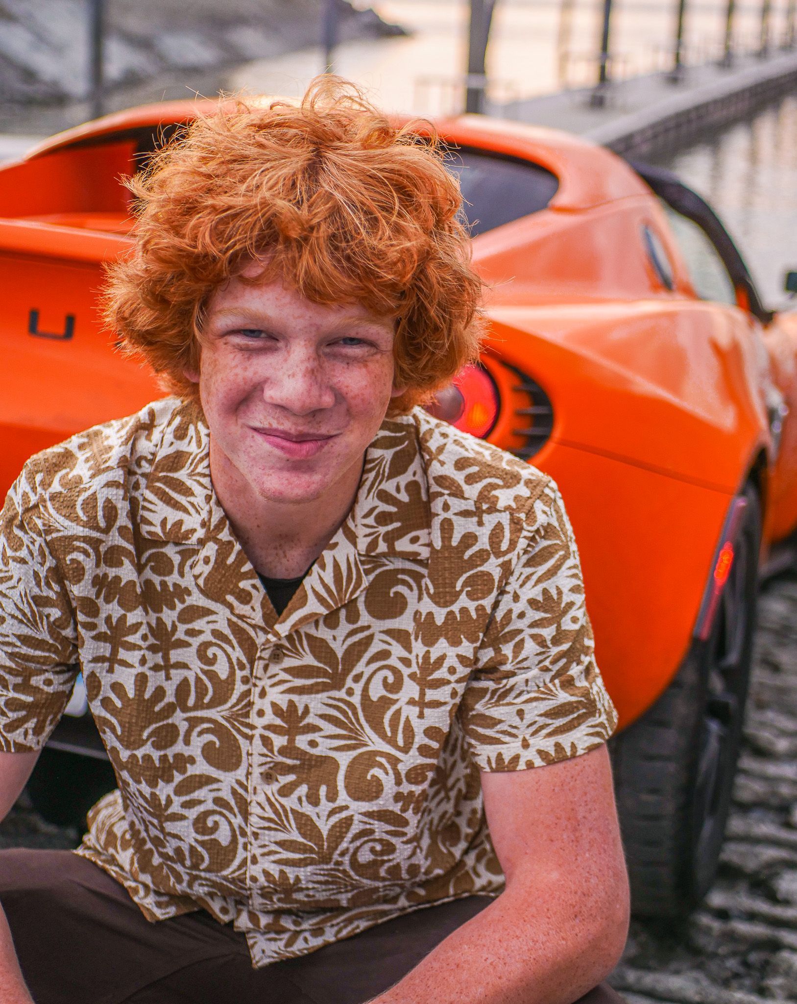 A young man with red hair is sitting in front of an orange car.