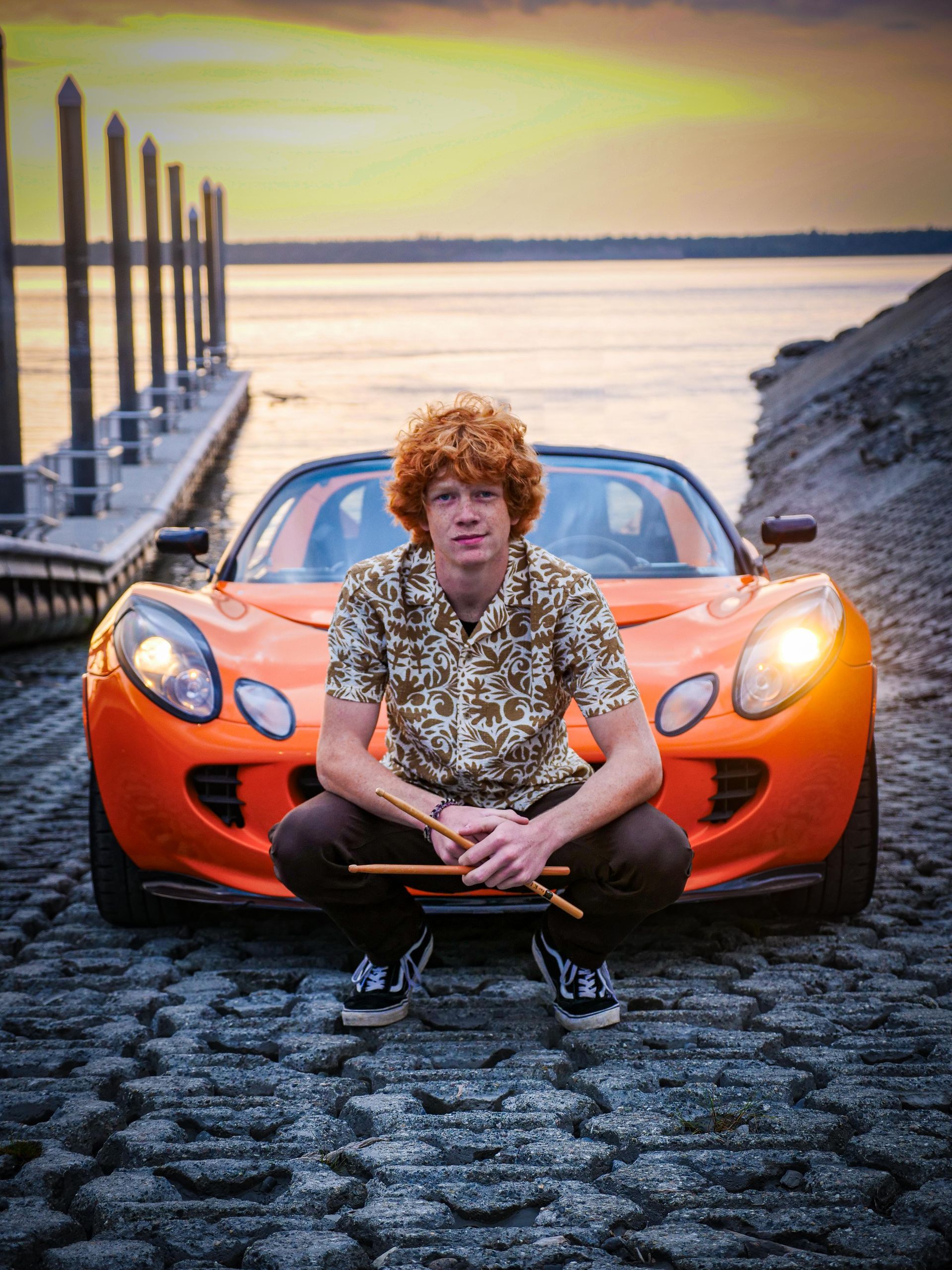 A man with red hair is kneeling in front of an orange sports car.