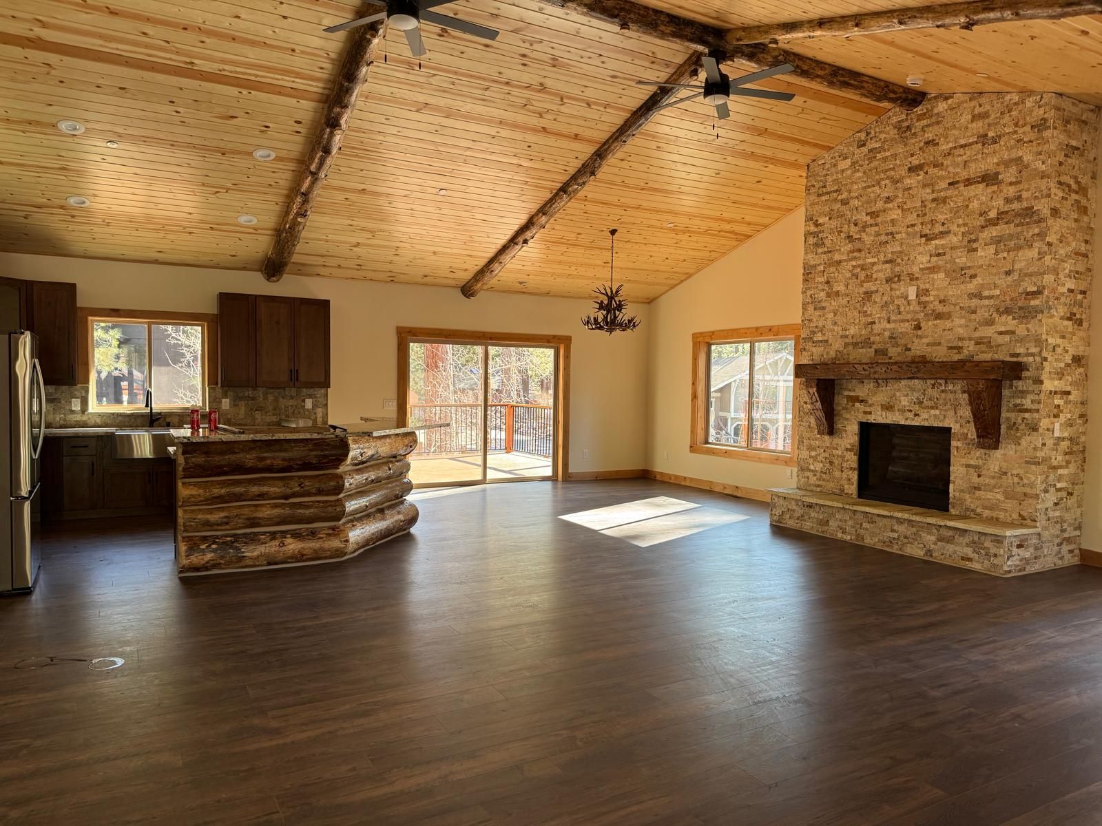 Spacious living area with wooden ceiling, stone fireplace, and dark flooring. Kitchen with wood cabinets.
