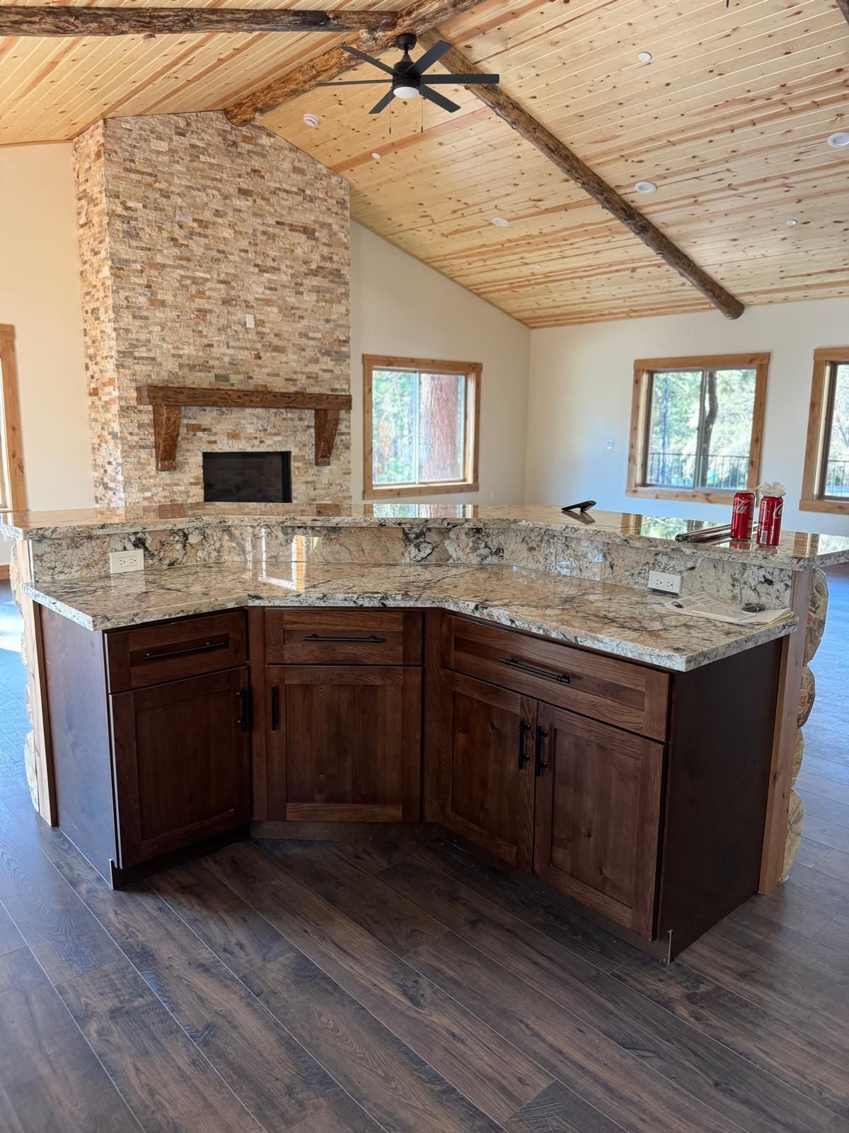 Rustic kitchen with granite countertops, dark cabinets, stone fireplace, and wood ceiling.