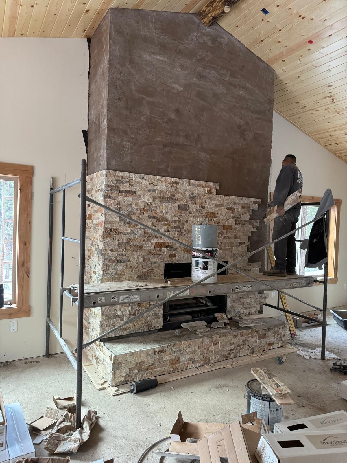 Man working on stone fireplace installation. Scaffolding, bucket, and unfinished walls present.