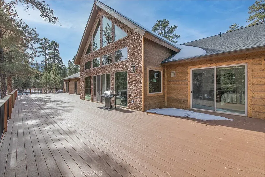 Wooden-sided house with a large deck, snow patches, and tall windows. Trees in the background under a blue sky.