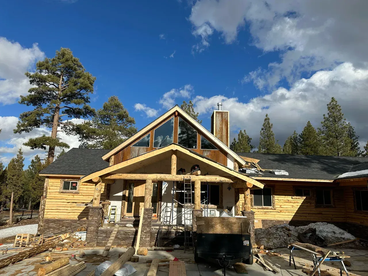 Log cabin under construction with exposed wooden logs against a blue sky, surrounded by pine trees.
