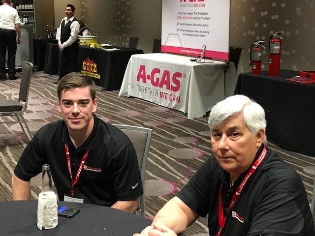 Two men are sitting at a table in front of a sign that says a-gas