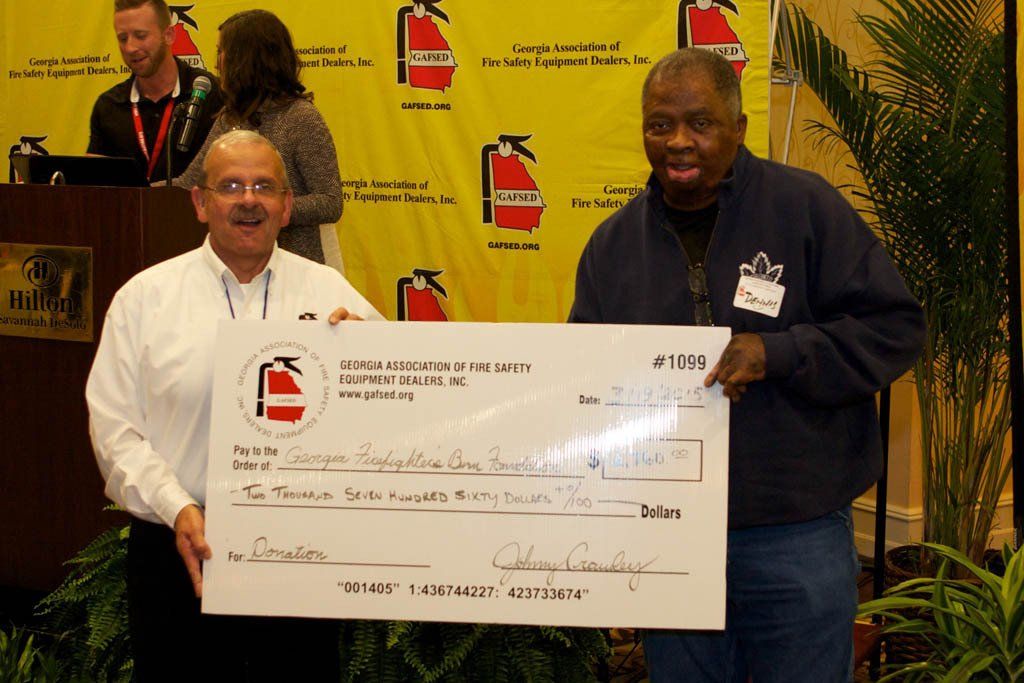 Two men holding a large check in front of a fire extinguisher wall