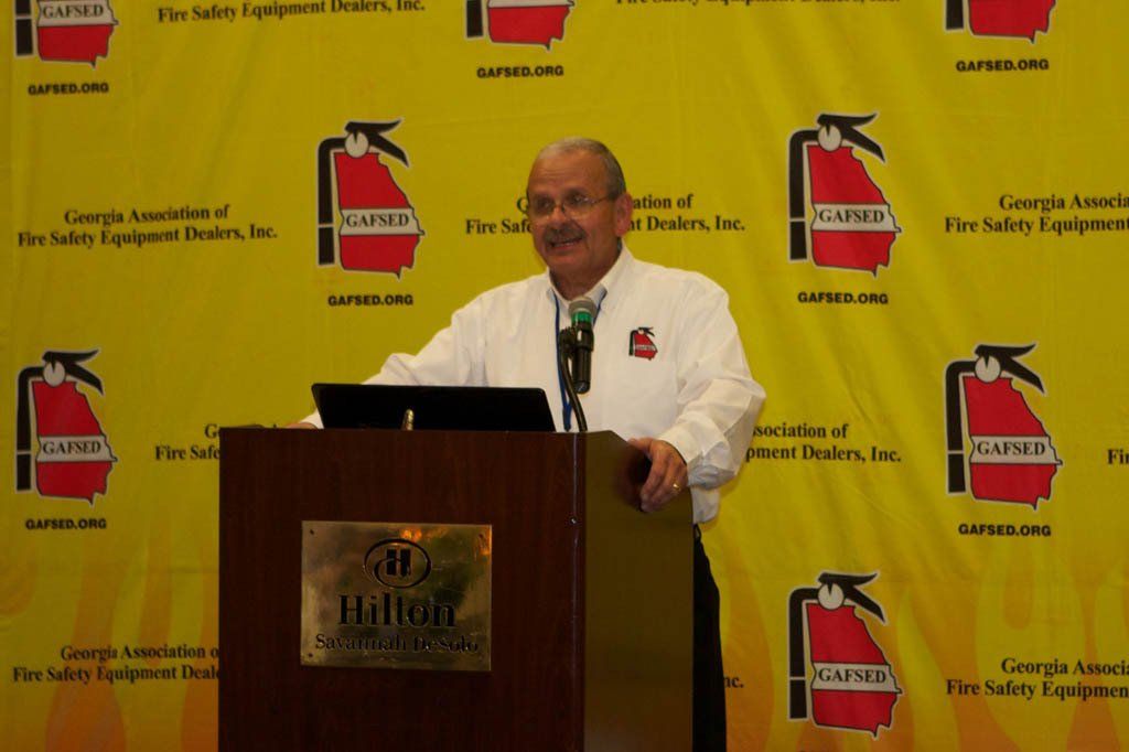 A man stands at a podium in front of a wall with fire extinguishers on it