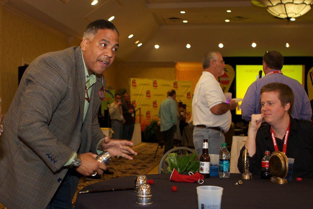 A man in a suit is standing in front of a table with two men sitting at it.