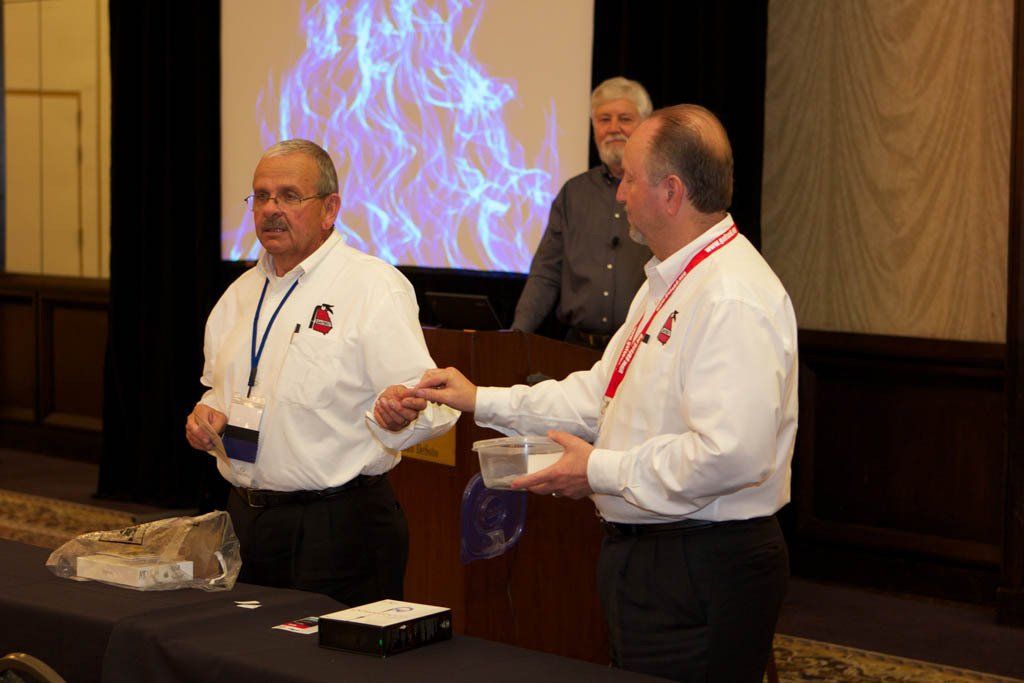 Two men are standing at a table in front of a projector screen with flames on it.