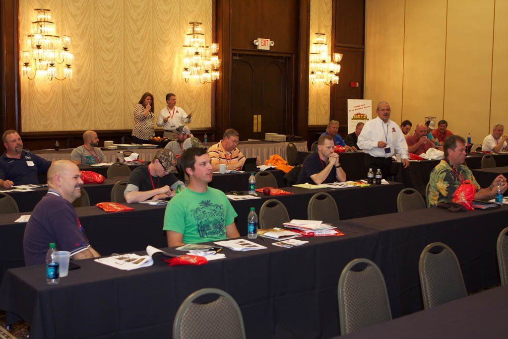A group of people are sitting at long tables in a conference room