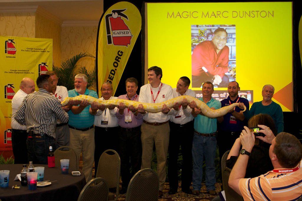 A group of men holding a snake in front of a screen that says magic marc dunston