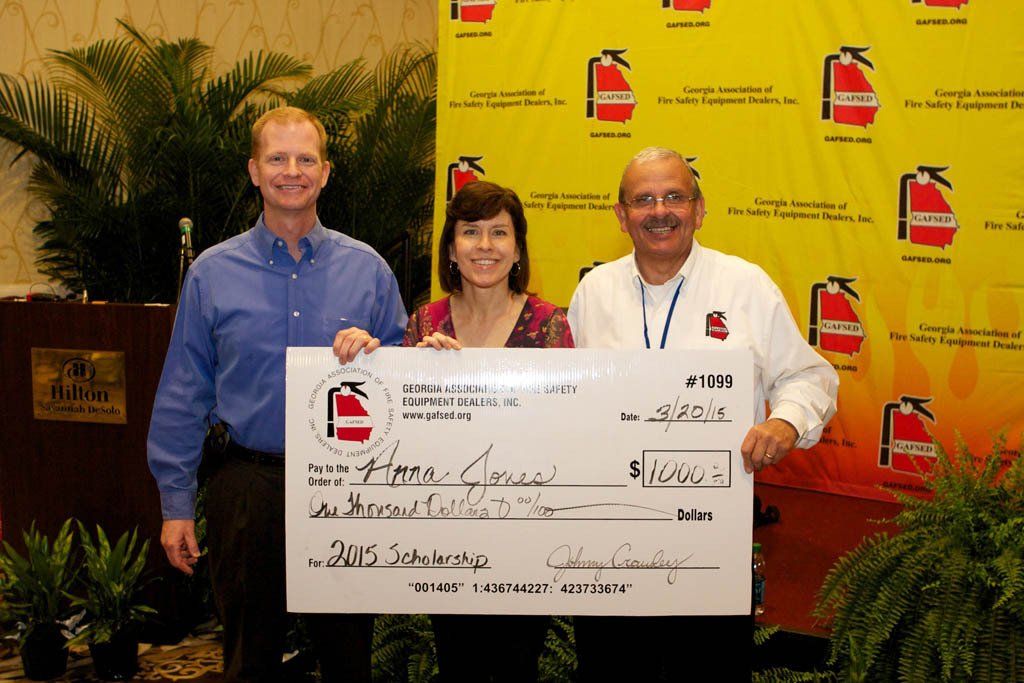 Two men and a woman holding a large check in front of a wall with fire extinguishers on it