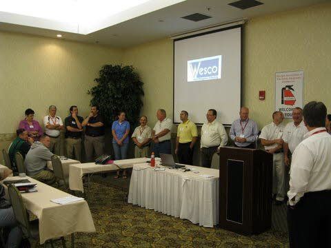 A group of people are standing around a podium in front of a projector screen that says wesco.